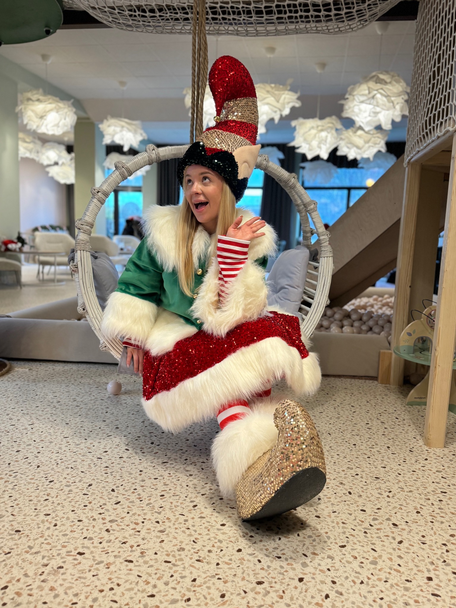 Uśmiechnięta animatorka w kostiumie elfa, siedząca na huśtawce w jasnym wnętrzu z dekoracyjnymi lampami. Widoczny fragment sali zabaw z kulkami.