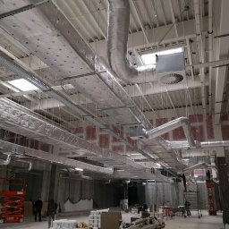 C&M Sp. z o.o. - Montaż systemu wentylacyjnego z izolacją termiczną w dużej hali, widoczna platforma robocza i pracownicy na tle surowych ścian.