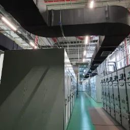 Długi, czarny kanał wentylacyjny biegnący pod sufitem hali przemysłowej, obok rzędów szaf sterowniczych i drabiny na ścianie.