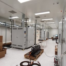 C&M Sp. z o.o. - Wnętrze sterylnego pomieszczenia z wentylacją, widoczne kanały wentylacyjne, urządzenia, skrzynie z materiałami i osoba pracująca przy komputerze w tle.