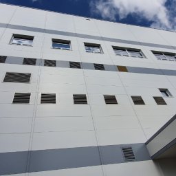 C&M Sp. z o.o. - Elewacja budynku przemysłowego z licznymi kratkami wentylacyjnymi, widoczna z dołu, z fragmentem żółtego kasku w prawym dolnym rogu.