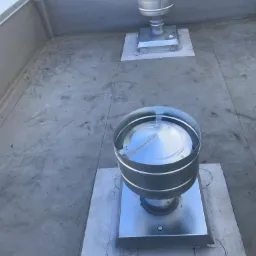 Dwa metalowe kominki wentylacyjne na płaskim, betonowym dachu z widocznymi śladami po montażu i zarysowanymi okręgami wokół podstawy, z fragmentem ulicy w tle.