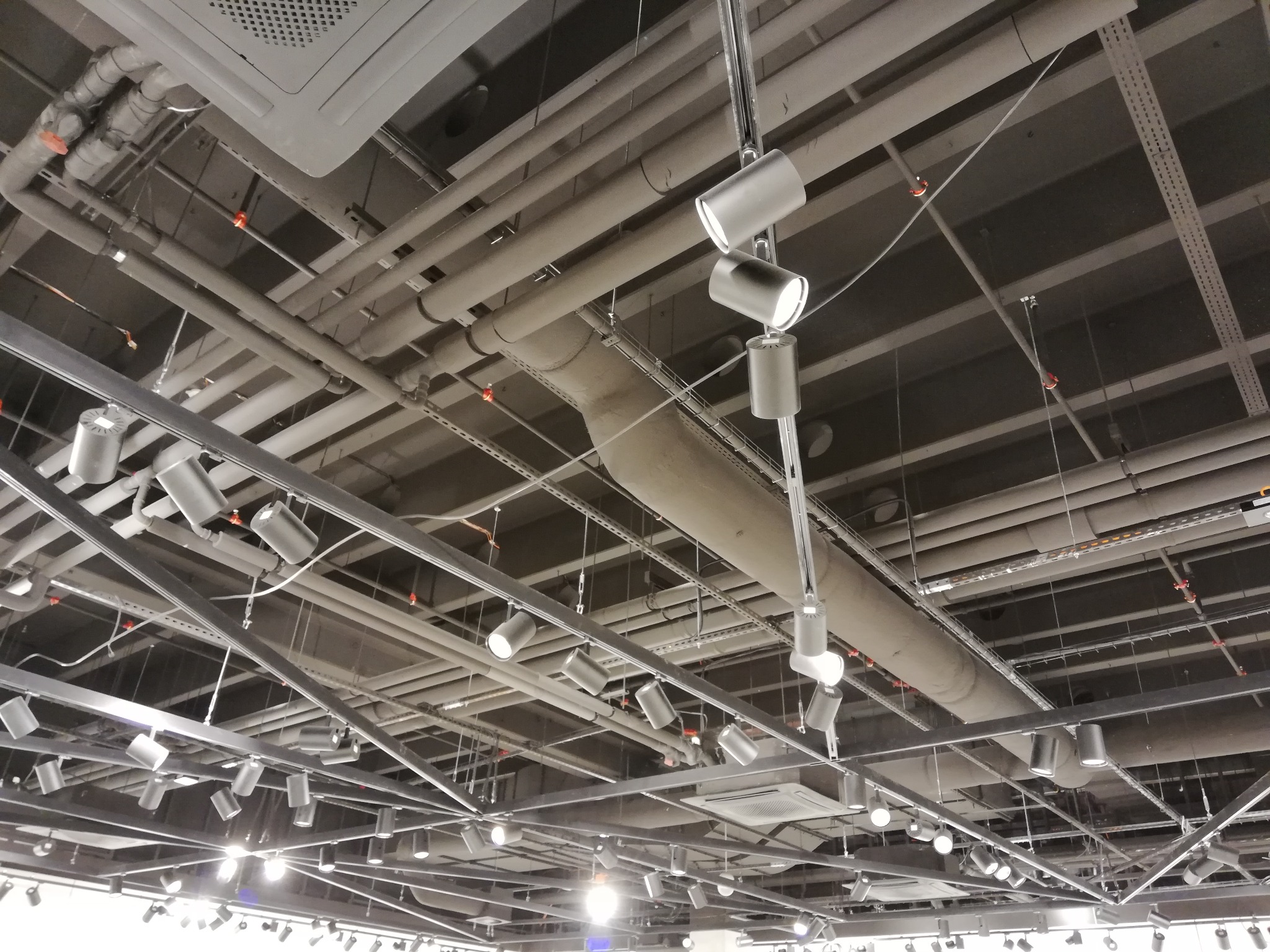 Skomplikowana sieć rur i przewodów pod sufitem, z licznymi punktowymi lampami oświetleniowymi, widoczne elementy instalacji przeciwpożarowej i wentylacyjnej.