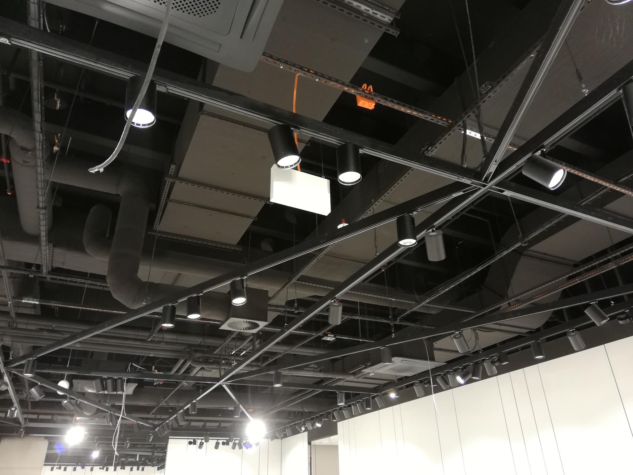 Skomplikowana instalacja pod sufitem z widocznymi rurami, przewodami i reflektorami punktowymi, tworząca industrialny wygląd wnętrza.
