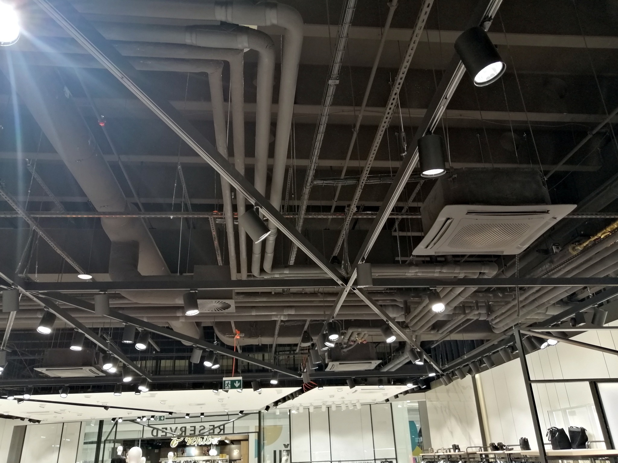 Widok na sufit w sklepie z widocznymi rurami wentylacyjnymi, przewodami i oświetleniem punktowym.