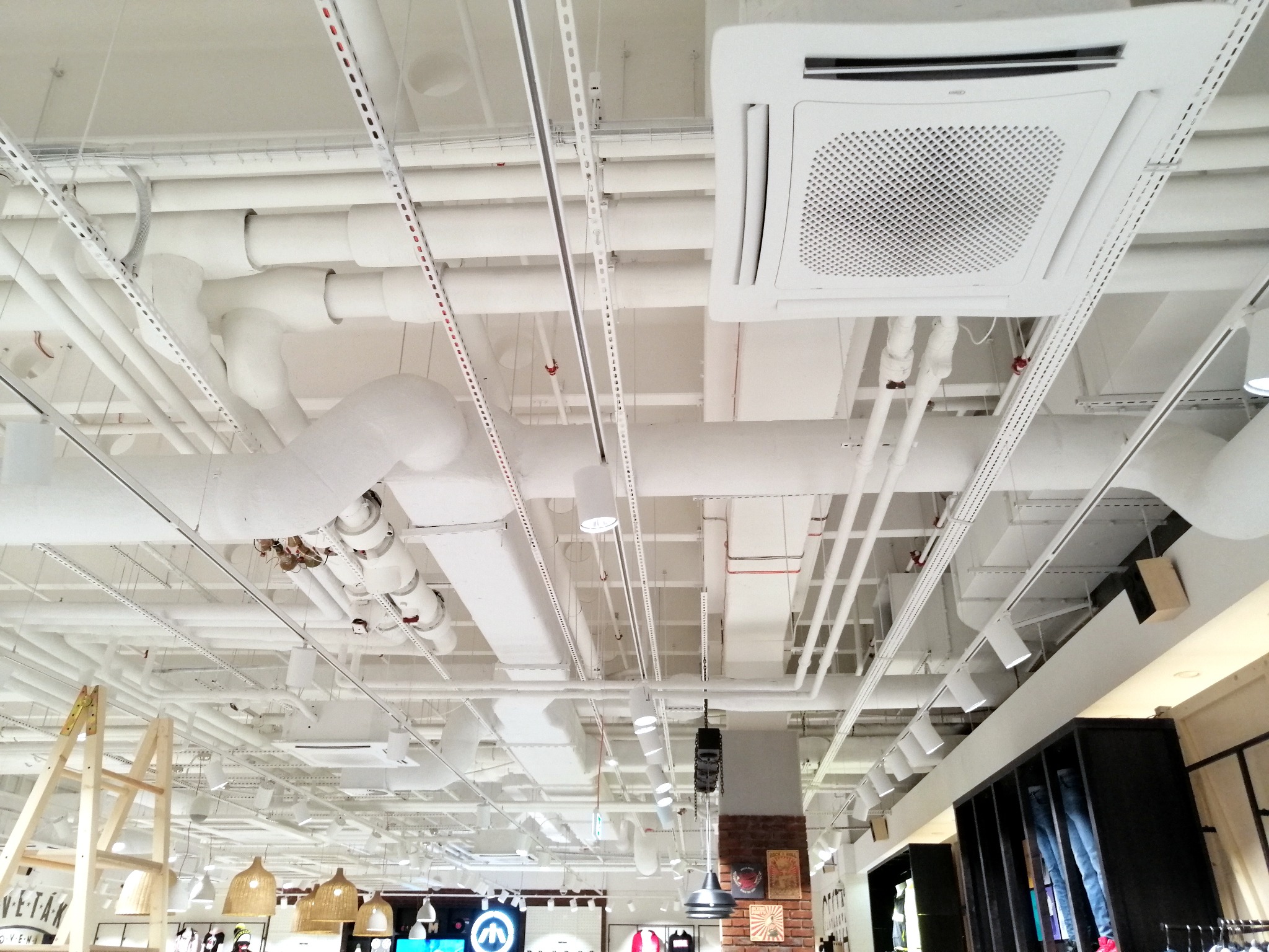 Biały sufit z widocznymi rurami, kanałami wentylacyjnymi, instalacjami i klimatyzatorem kasetonowym w pomieszczeniu handlowym.