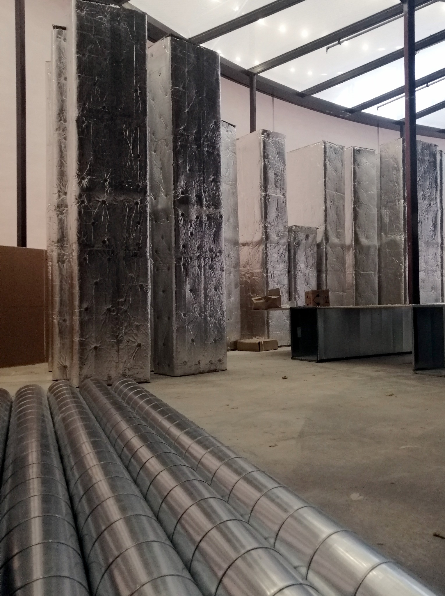 Izolowane kanały wentylacyjne, pionowe i okrągłe, leżące na podłodze hali produkcyjnej przed montażem.