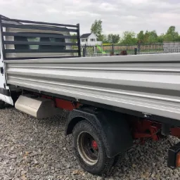 Biały samochód dostawczy z otwartą, pustą skrzynią ładunkową, zaparkowany na żwirowym podłożu, z widocznym domem w tle.