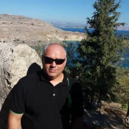 Mężczyzna w czarnej koszulce polo z zielonymi akcentami i okularach przeciwsłonecznych pozuje na tle skalistego krajobrazu z widokiem na morze i drzewa.