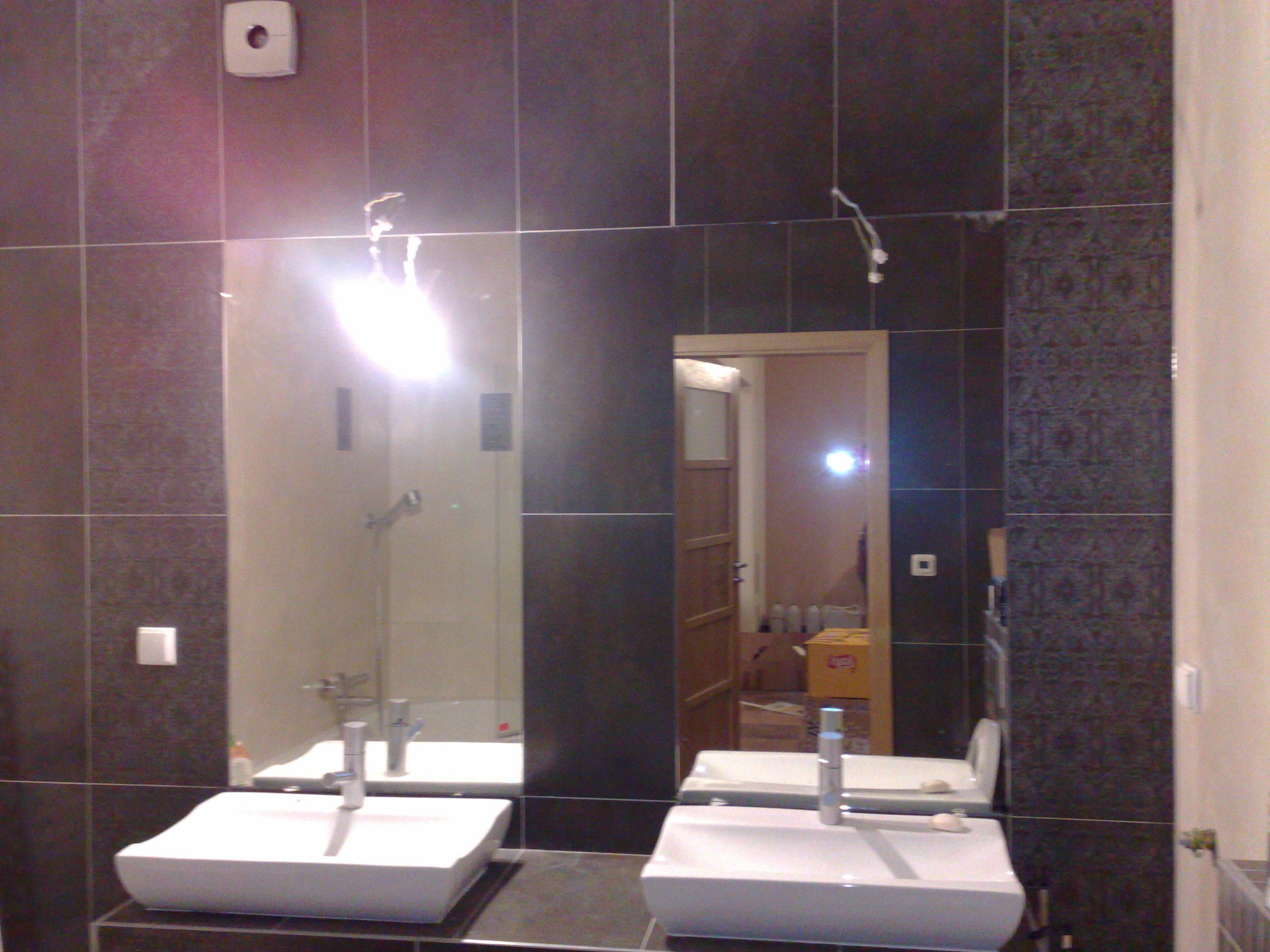 Nowoczesna łazienka z ciemnymi, błyszczącymi płytkami, podwójną umywalką z minimalistyczną armaturą i dużym lustrem odbijającym kabinę prysznicową; widoczne niedokończone instalacje elektryczne.