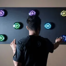 Mężczyzna tyłem dotyka dłońmi interaktywnego panelu z okrągłymi, podświetlanymi punktami w kolorach fioletowym, zielonym, żółtym i niebieskim, prawdopodobnie w celach edukacyjnych lub terapeutycznych.