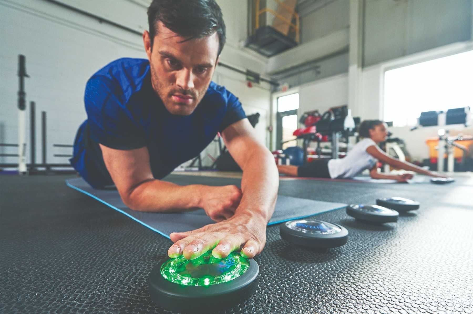 Mężczyzna w pozycji plank na macie, dotyka dłonią interaktywnego dysku z zielonym podświetleniem LED w siłowni, w tle ćwicząca kobieta.