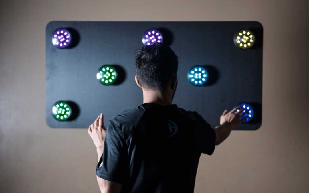 Mężczyzna tyłem dotyka dłońmi interaktywnego panelu z okrągłymi, podświetlanymi punktami w kolorach fioletowym, zielonym, żółtym i niebieskim, prawdopodobnie w celach edukacyjnych lub terapeutycznych.