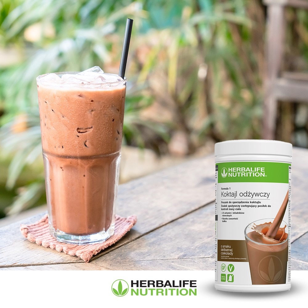 Mrożony koktajl czekoladowy w wysokiej szklance z czarną słomką, obok opakowanie proszku Herbalife Formula 1 Koktajl Odżywczy o smaku delikatnej czekolady, na drewnianym stole z widocznym logo...