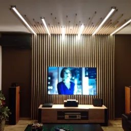 Nowoczesny salon z telewizorem, drewnianą ścianą z pionowych listew i oświetleniem LED zamontowanym na suficie, tworzącym dekoracyjny wzór.