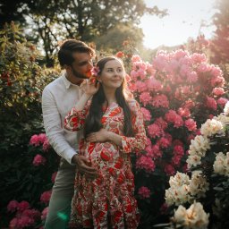 Sylwia Klatka Fotografia - Uśmiechnięta, ciężarna kobieta w kwiecistej sukience, obejmowana przez mężczyznę w ogrodzie pełnym różowych rododendronów w słoneczny dzień.