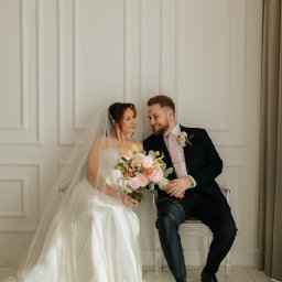 Sylwia Klatka Fotografia - Uśmiechnięta para młoda siedzi na krzesłach na tle białej ściany z dekoracyjnymi panelami. Panna młoda trzyma bukiet różowych kwiatów.