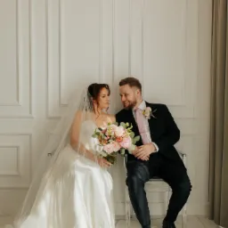 Uśmiechnięta para młoda siedzi na krzesłach na tle białej ściany z dekoracyjnymi panelami. Panna młoda trzyma bukiet różowych kwiatów.