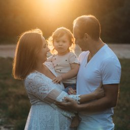 Sylwia Klatka Fotografia - Ciepłe ujęcie rodziny w plenerze: kobieta w koronkowej sukience trzyma dziecko, obok stoi mężczyzna. Złote światło zachodzącego słońca dodaje uroku.