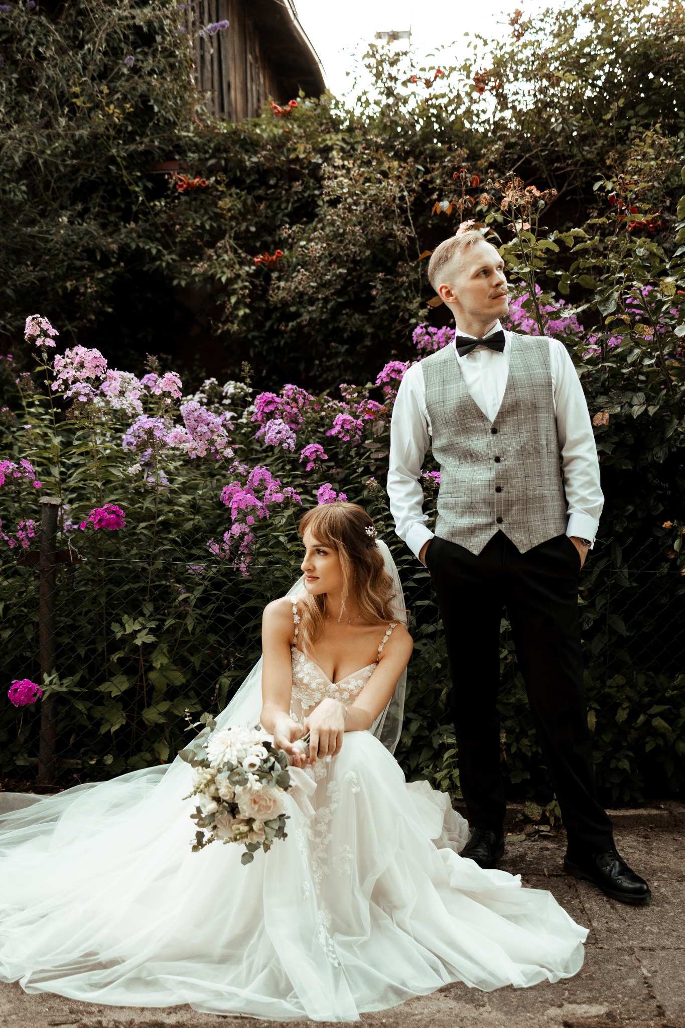 Panna młoda w koronkowej sukni siedzi z bukietem na tle kwitnących floksów, obok stoi pan młody w kraciastej kamizelce i muszce.