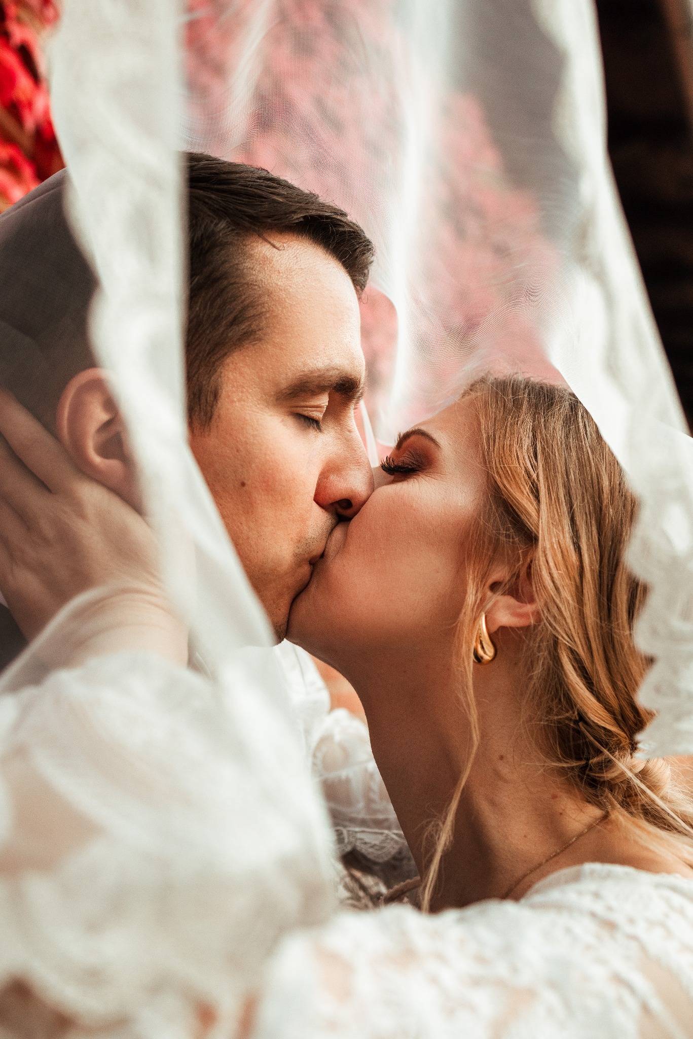 Młoda para całuje się pod delikatnym, koronkowym welonem, złote kolczyki panny młodej błyszczą w ciepłym świetle.