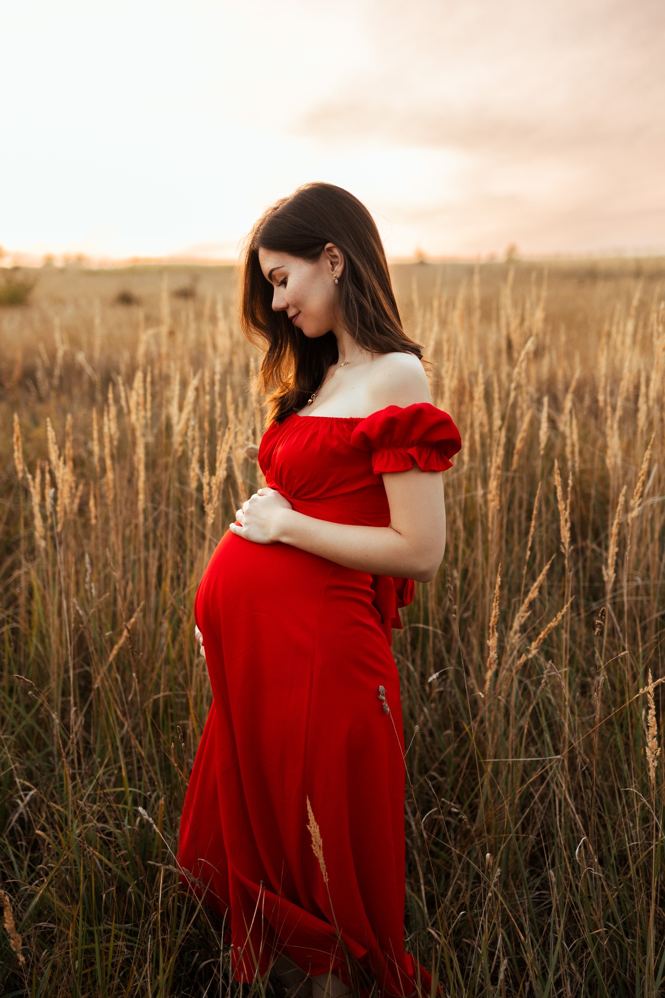Kobieta w czerwonej sukni ciążowej stoi na polu, trzymając dłonie na brzuchu, z pochyloną głową i delikatnym uśmiechem.