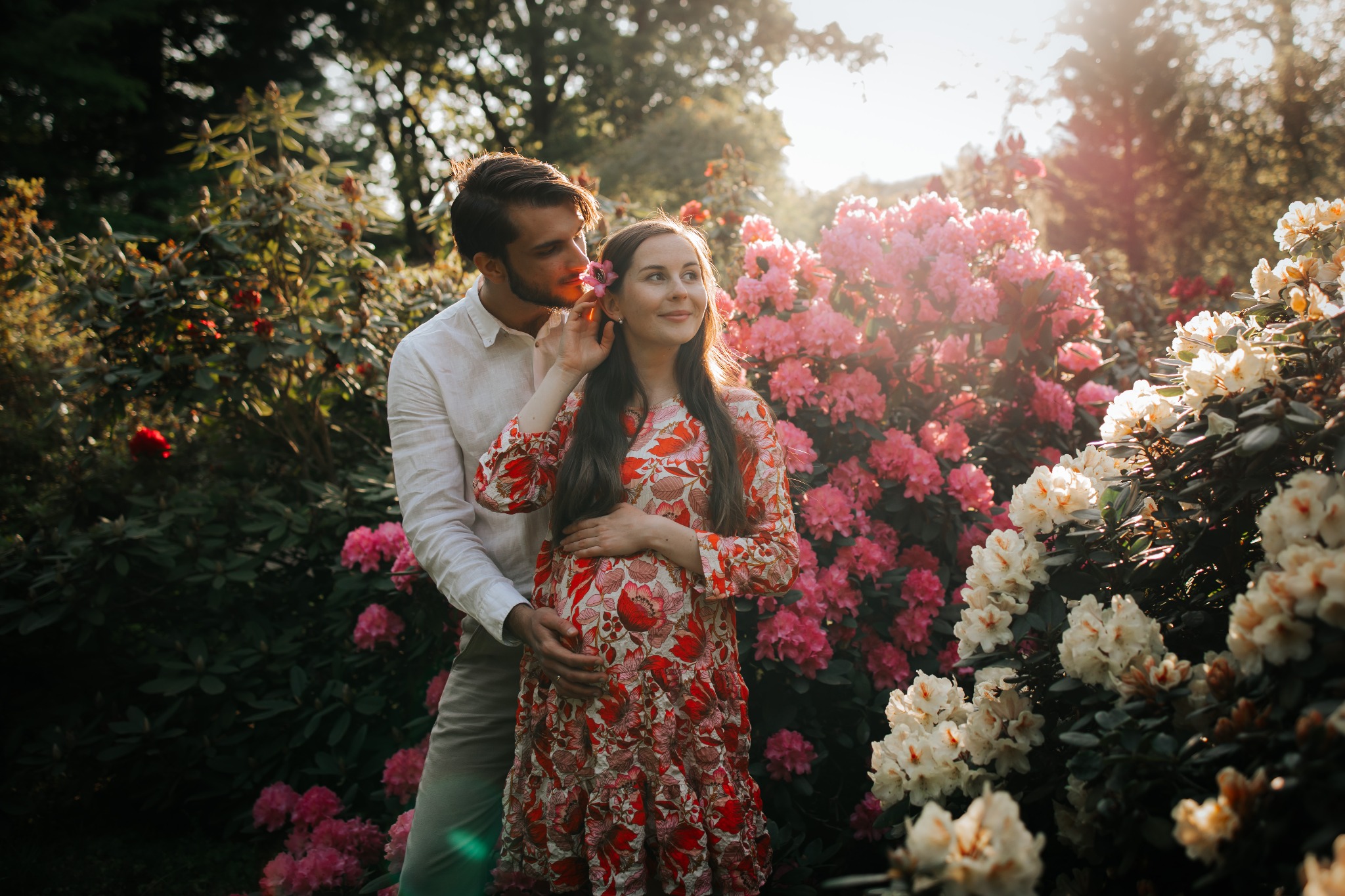 Uśmiechnięta, ciężarna kobieta w kwiecistej sukience, obejmowana przez mężczyznę w ogrodzie pełnym różowych rododendronów w słoneczny dzień.