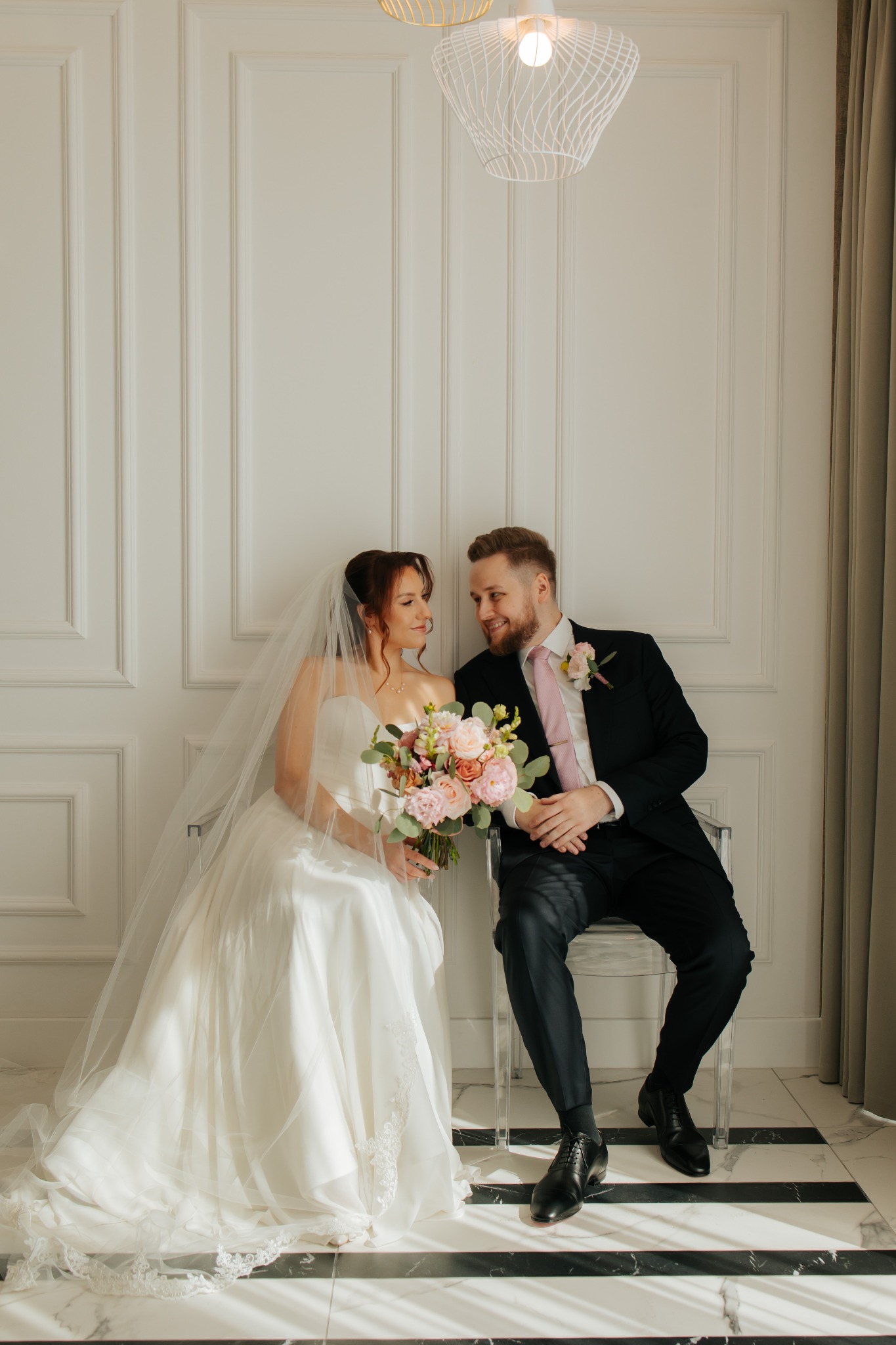 Uśmiechnięta para młoda siedzi na krzesłach na tle białej ściany z dekoracyjnymi panelami. Panna młoda trzyma bukiet różowych kwiatów.