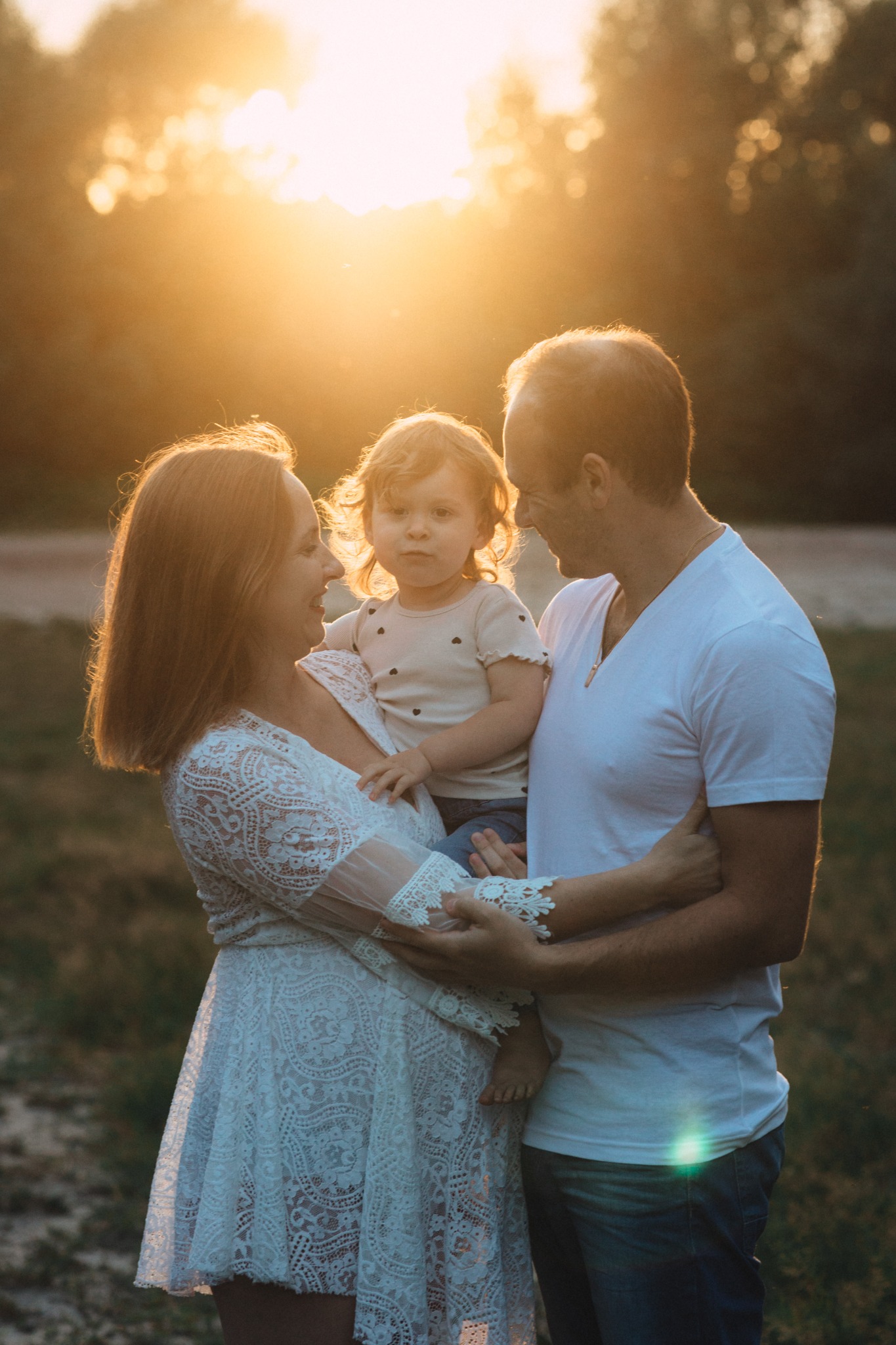 Ciepłe ujęcie rodziny w plenerze: kobieta w koronkowej sukience trzyma dziecko, obok stoi mężczyzna. Złote światło zachodzącego słońca dodaje uroku.