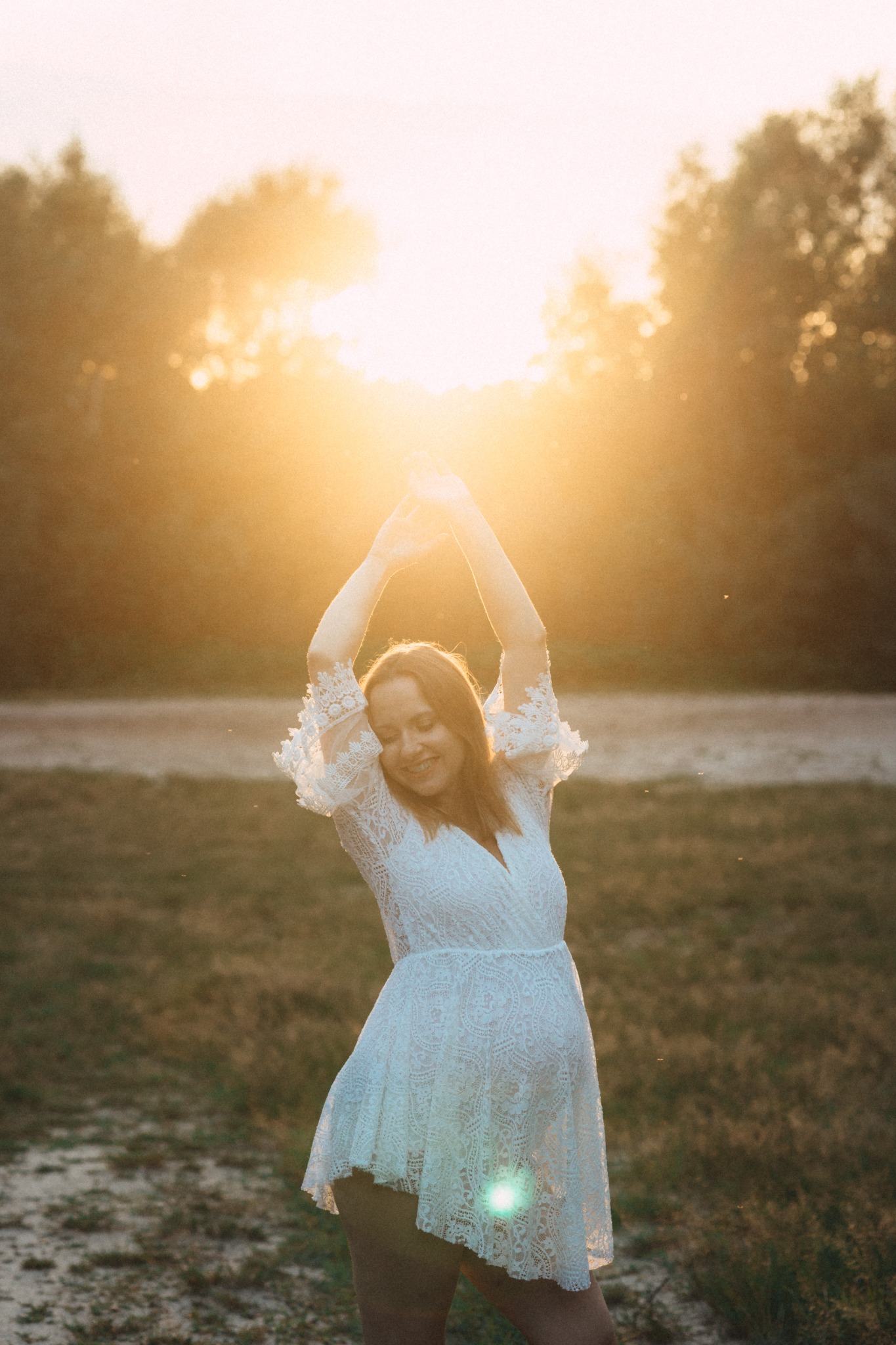 Uśmiechnięta kobieta w koronkowej sukience, z uniesionymi rękoma, na tle zachodzącego słońca i drzew. Romantyczna sesja w plenerze.