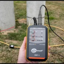 Miernik Sonel ERP-1 w dłoni, z żółtym przewodem okalającym betonowy słup, prawdopodobnie podczas pomiarów uziemień.
