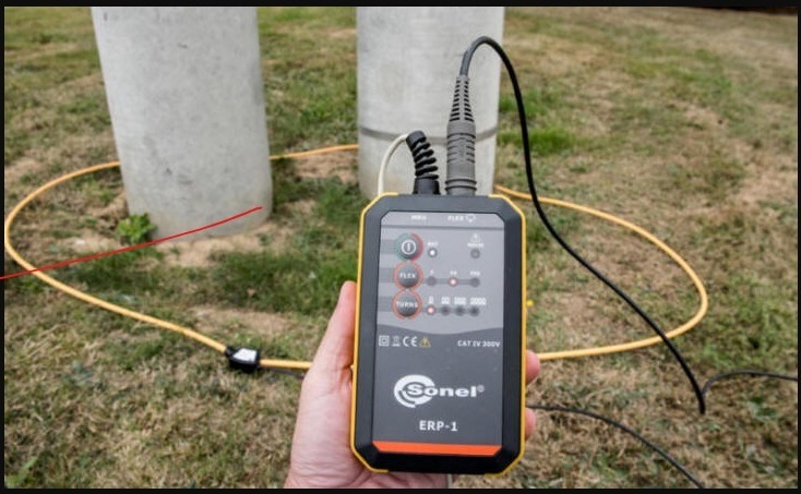 Miernik Sonel ERP-1 w dłoni, z żółtym przewodem okalającym betonowy słup, prawdopodobnie podczas pomiarów uziemień.