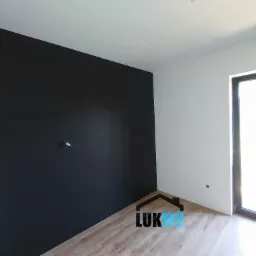 Wnętrze pokoju po remoncie: jedna ściana pomalowana na czarno, pozostałe na biało, widoczna podłoga laminowana i okno. Na ścianie widoczny punkt instalacji elektrycznej.