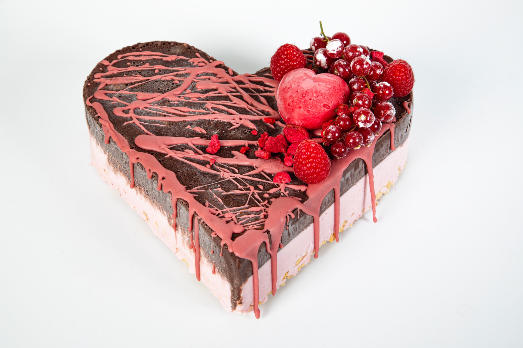 Tort w kształcie serca z ciemną polewą, różową glazurą i dekoracją z malin, porzeczek oraz małego serca z galaretki, na białym tle.