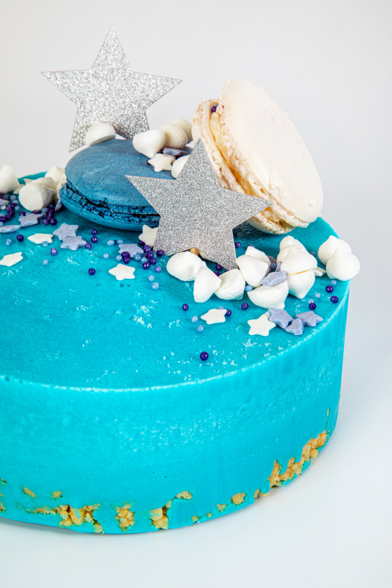 Turkusowy tort z błyszczącymi gwiazdkami, makaronikami i posypką w kształcie gwiazd i kulek na białym tle.