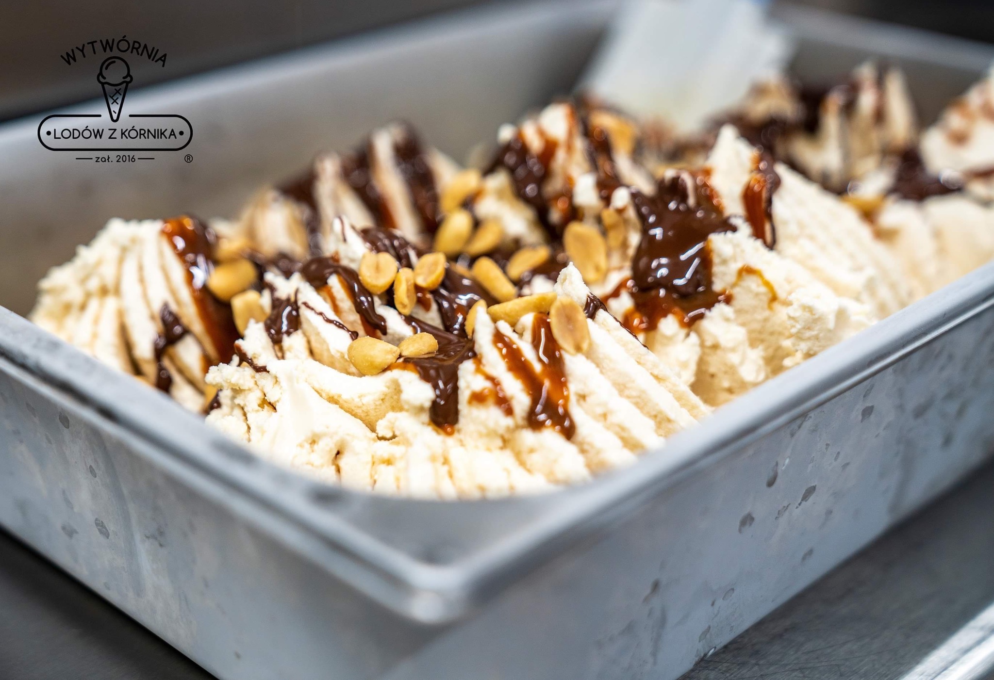 Metalowa kuweta z kremowymi lodami polanymi karmelem i czekoladą, posypana orzeszkami ziemnymi, widoczne logo producenta lodów.