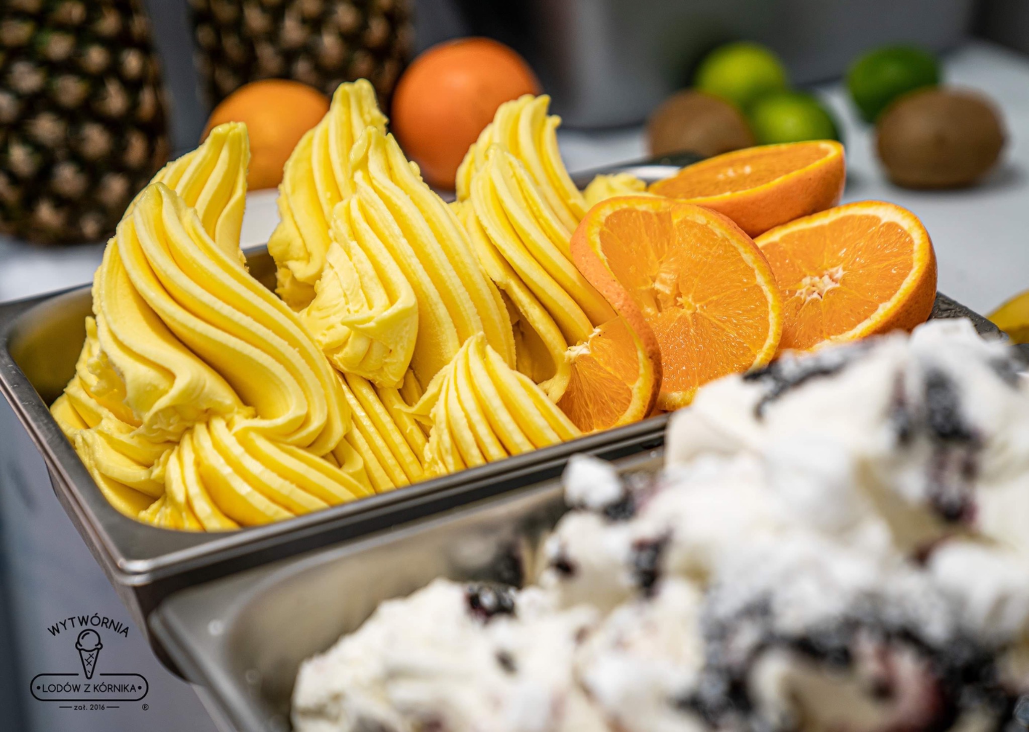 Żółte lody o smaku mango w metalowej kuwecie, udekorowane plasterkami pomarańczy, z ananasami i kiwi w tle, obok lody śmietankowe z jagodami.