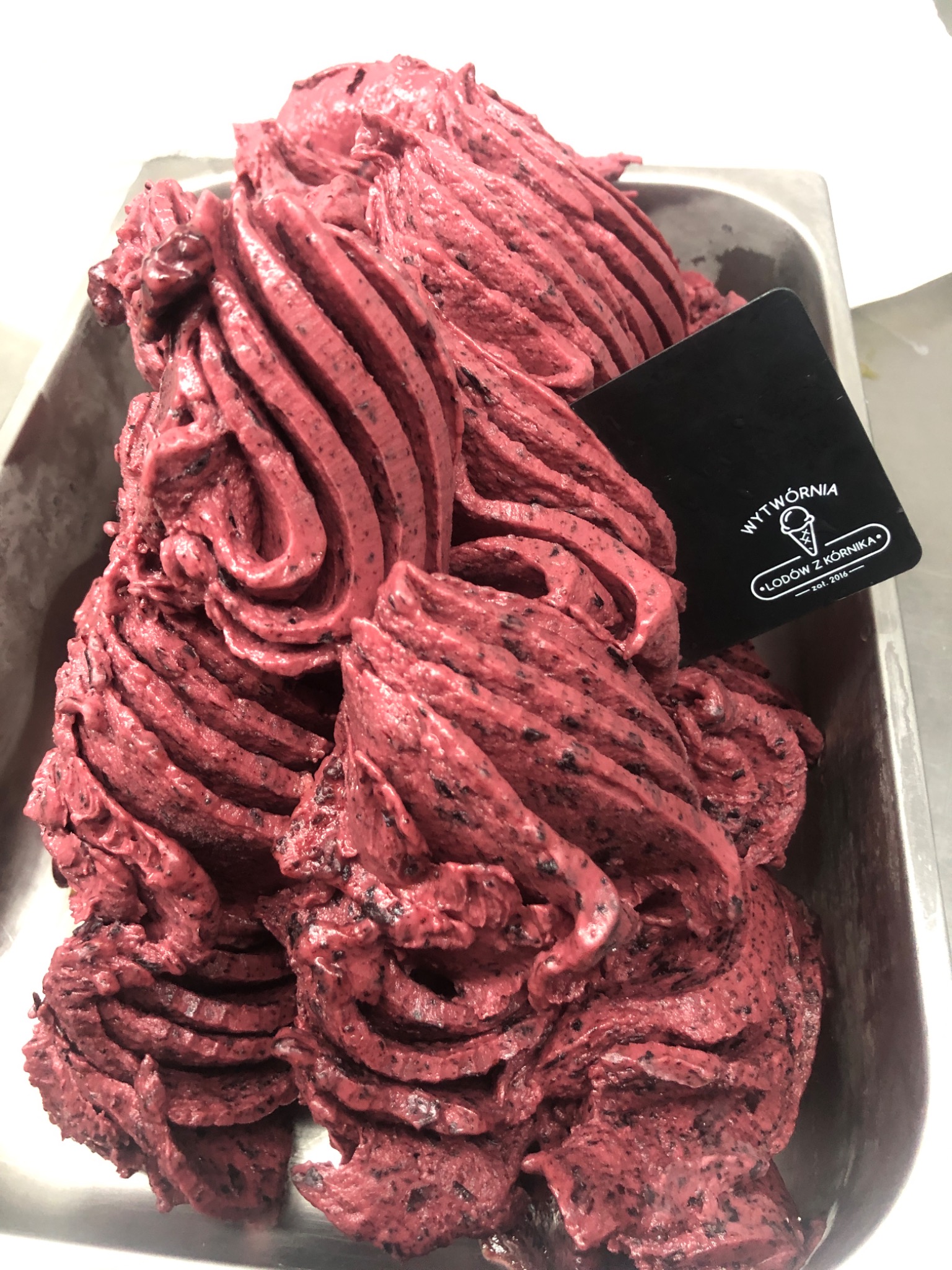 Porcja lodów jagodowych w metalowej kuwecie, z czarną etykietą producenta 'Wytwórnia Lodów z Kórnika'.