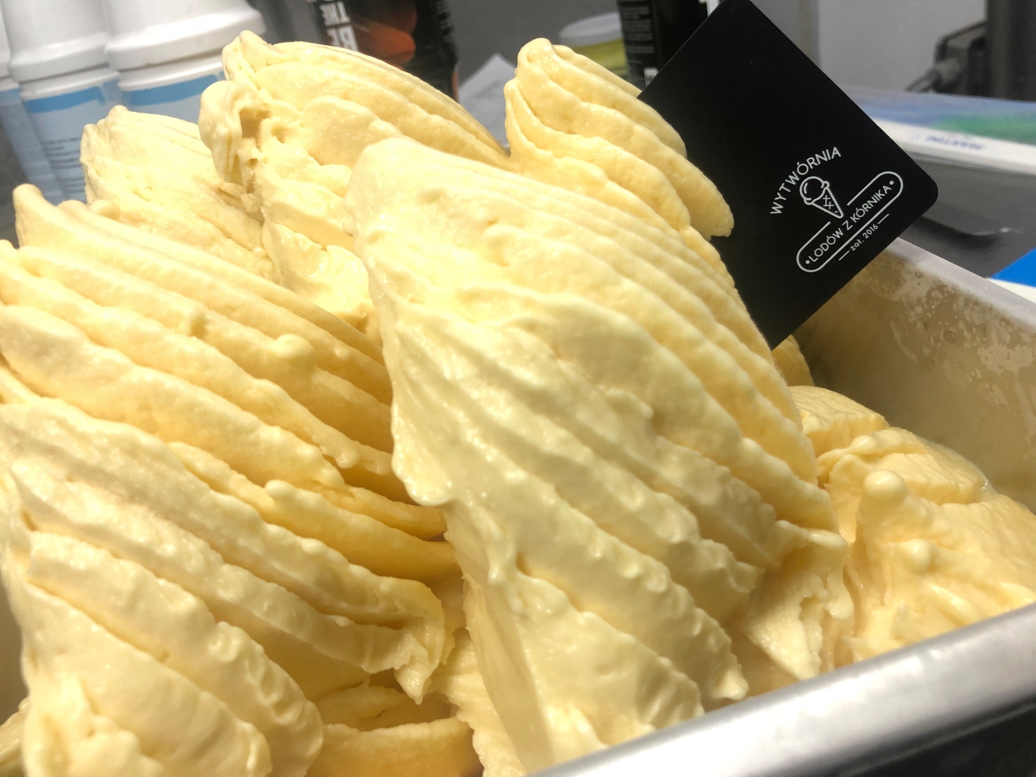 Waniliowe lody włoskie ułożone w metalowej kuwecie, obok czarna tabliczka z logo lodziarni.