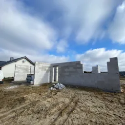 Wznoszone ściany parterowego domu z betonu komórkowego, widoczne palety z bloczkami i fragment sąsiedniego budynku.