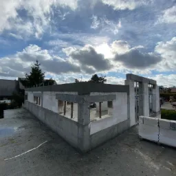 Surowy stan budynku z betonu komórkowego, widoczne zbrojenia w nadprożach okiennych, fundament wylany, paleta z pustakami na dachu, błękitne niebo z chmurami w tle.