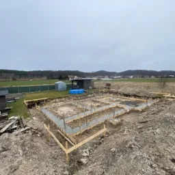 Zalane betonem szalunki fundamentowe na placu budowy z widokiem na okoliczne pola i zabudowania w pochmurny dzień.