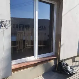 Białe okno przesuwne, widok z zewnątrz, mur w trakcie wykończenia, na parapecie ceglana okładzina, obok okna oparta miotła i czarny worek na śmieci.