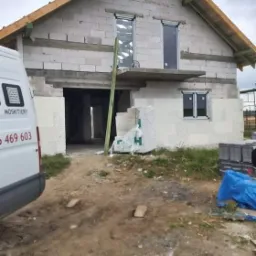 Budynek w trakcie budowy z zamontowanymi oknami i roletami zewnętrznymi; na pierwszym planie widoczny samochód dostawczy z napisem 'Moskitiery'.