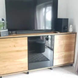 Drewniana szafka RTV z czarnymi ramami i lustrzanym frontem, na której stoi telewizor, głośnik i roślina doniczkowa, widoczna w salonie z szarą podłogą i zasłonami.