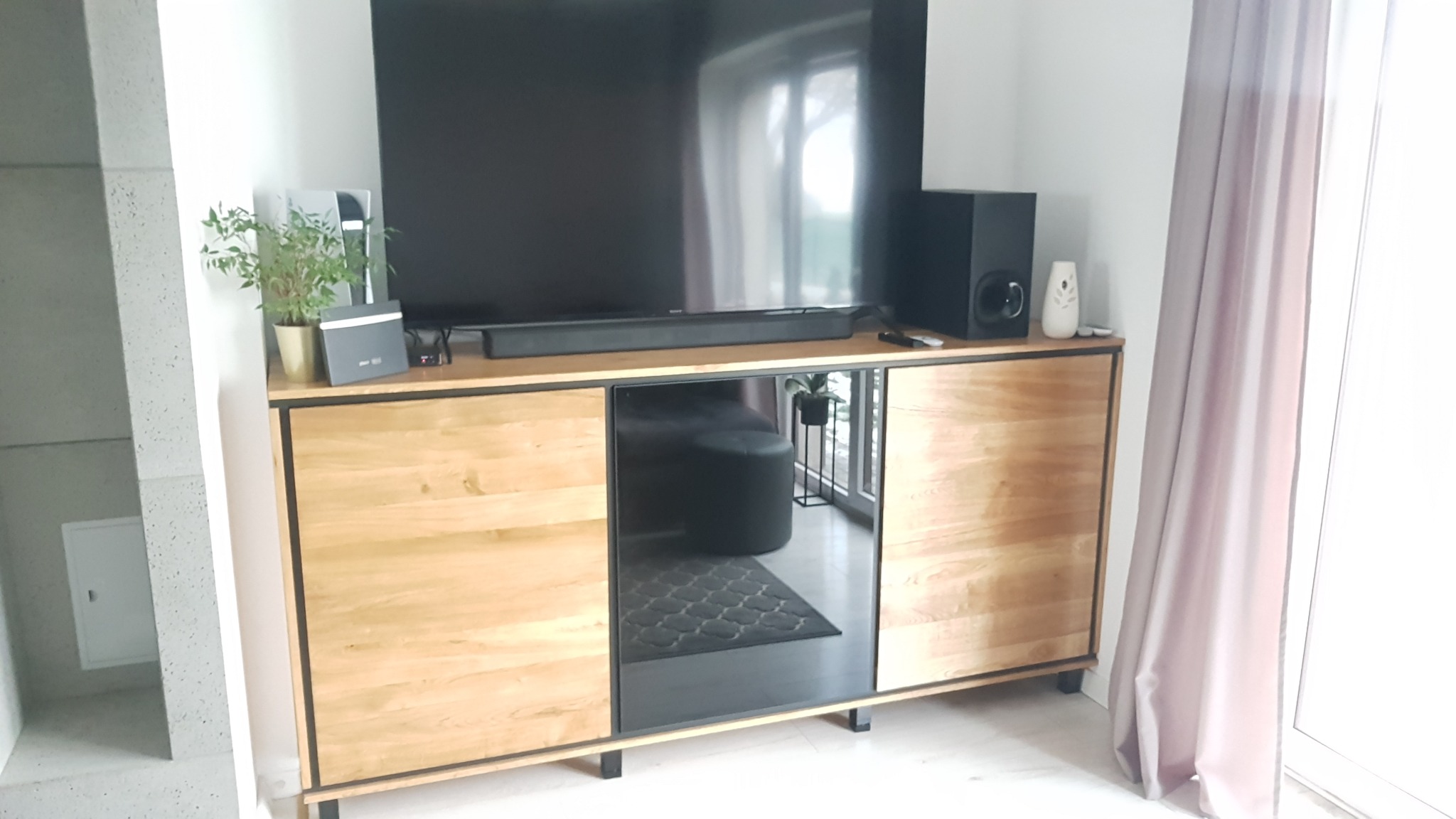 Drewniana szafka RTV z czarnymi ramami i lustrzanym frontem, na której stoi telewizor, głośnik i roślina doniczkowa, widoczna w salonie z szarą podłogą i zasłonami.