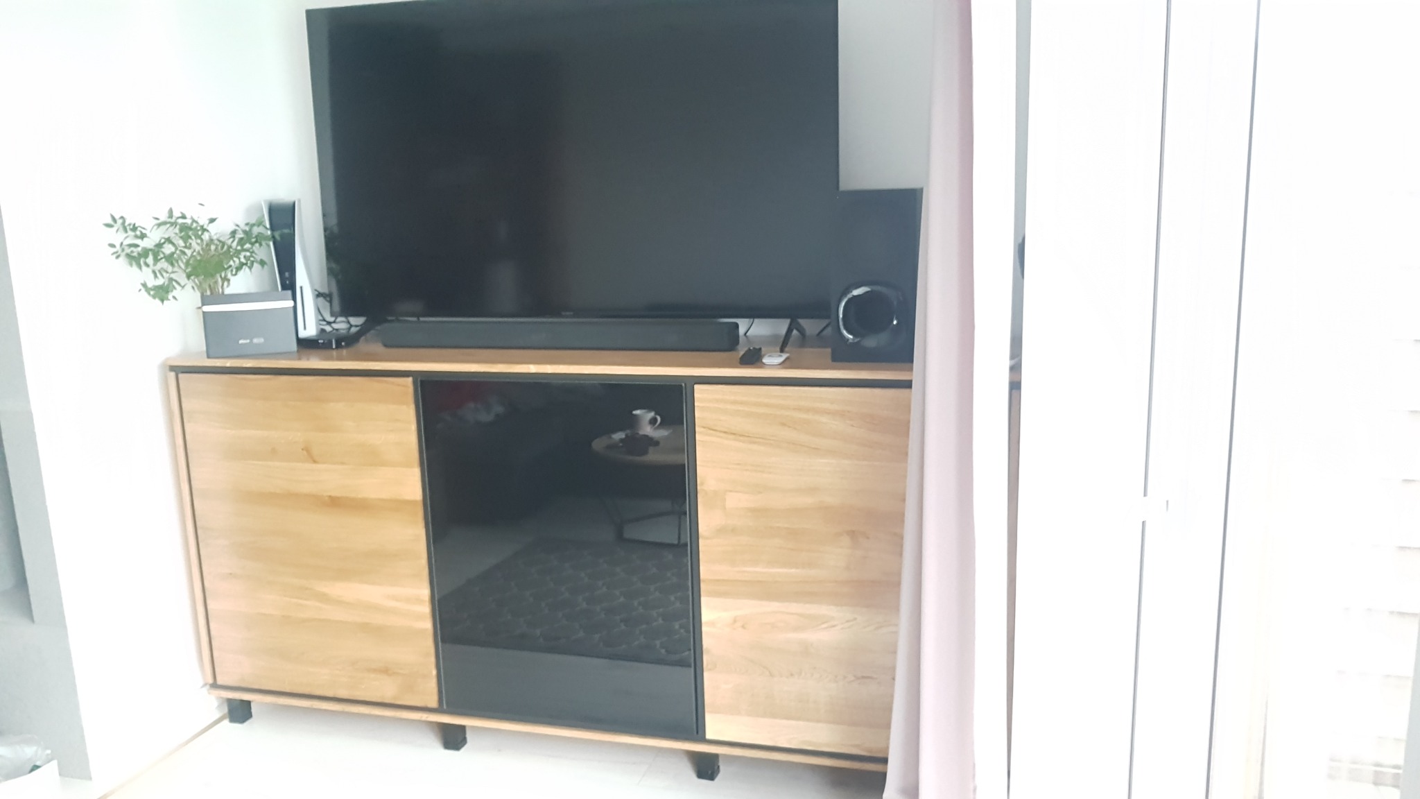 Nowoczesna szafka RTV z jasnego drewna z czarnymi, szklanymi frontami, na której stoi duży telewizor, soundbar, konsola PlayStation 5 i głośnik, z widocznym odbiciem wnętrza pokoju w szklanych...