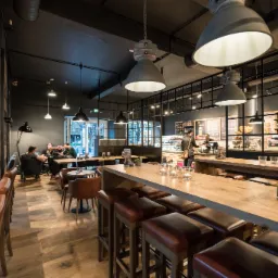 Wnętrze kawiarni z industrialnymi lampami wiszącymi nad drewnianym barem i skórzanymi stołkami, widoczne osoby siedzące przy stolikach w tle.