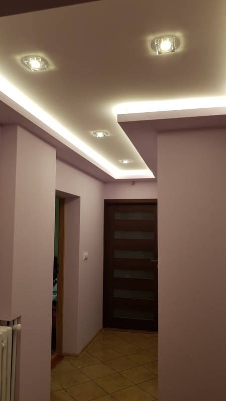 Korytarz z podwieszonym sufitem z oświetleniem LED, dodatkowo punktowe lampy sufitowe, ściany w odcieniach fioletu, brązowe drzwi z przeszkleniami, podłoga wyłożona płytkami w odcieniach brązu.