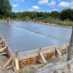 Wylewka fundamentowa, szalunek z desek, pracownik wyrównuje powierzchnię betonu, widoczne pomarańczowe pachołki i toaleta przenośna w tle.